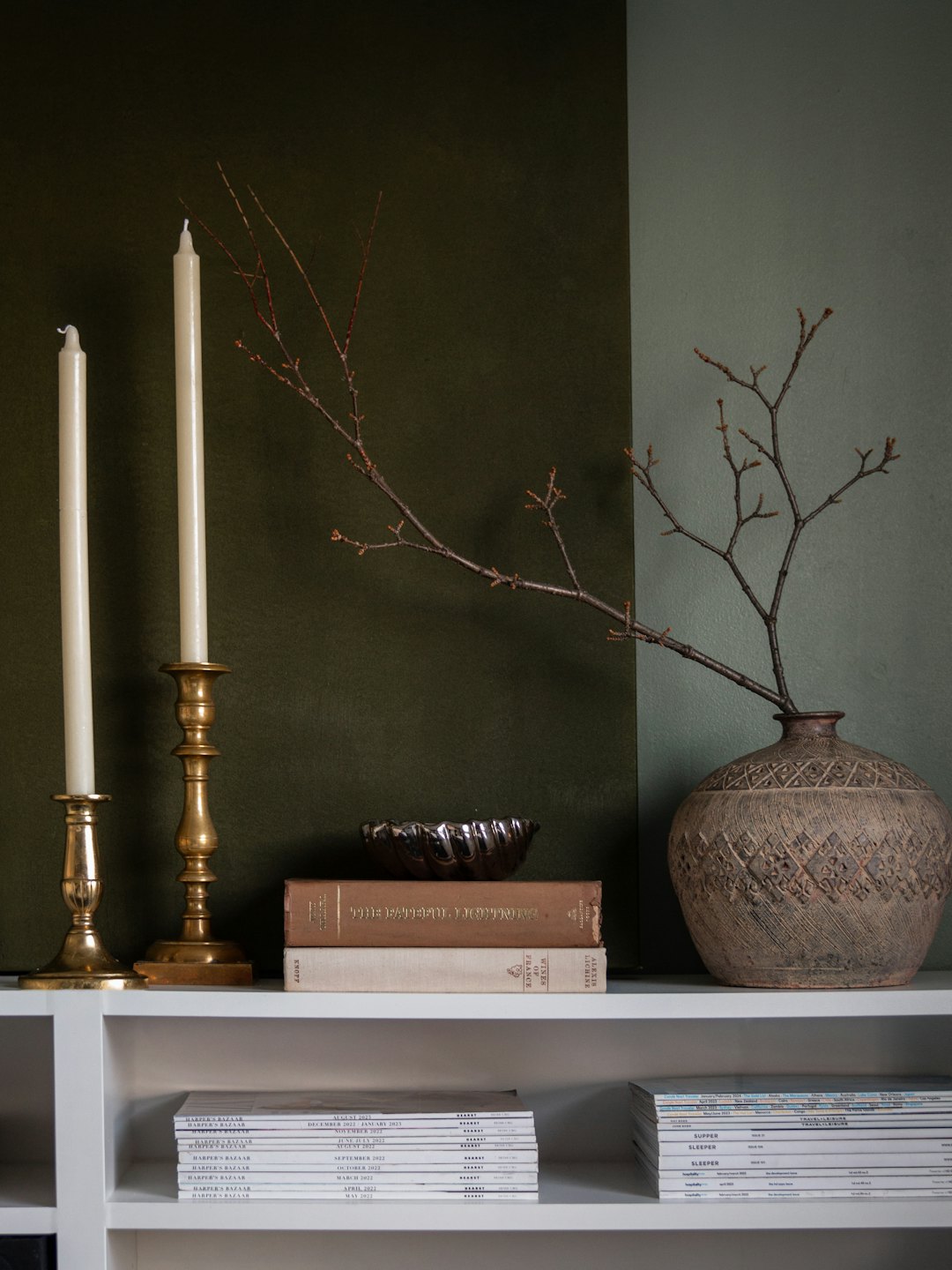 Decorated shelf with candles and books against a green wall.